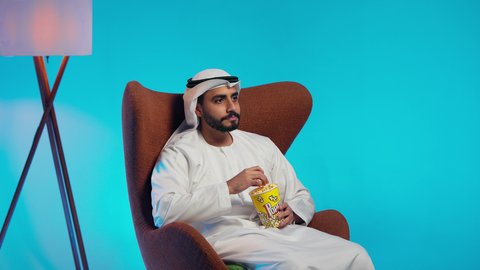 القيام بنشاطات ترفيهيه ممتعه، قضاء وقت الفراغ بمتابعة الافلام و المسلسلات المسلية، تناول سناك صحي و خفيف ، رجل عربي خليجي اماراتي يرتدي الكندورة والغترة يجلس على كرسي مريح ويقوم بتناول الفشار