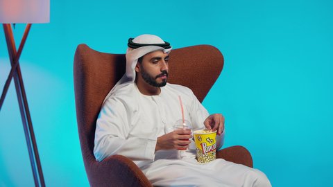قضاء وقت الفراغ بمتابعة الافلام و المسلسلات المسلية، تناول سناك صحي و خفيف ، رجل عربي خليجي اماراتي يرتدي الكندورة والغترة يجلس على كرسي مريح ويقوم بتناول الفشار