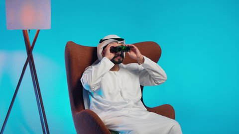 رجل عربي خليجي اماراتي يرتدي الكندورة والغتره يجلس على كرسي مريح يحمل بيده المنظار ثنائي العينين وينظر الى شيء ما، حب الاستكشاف والمغامرة، استعمال المنظار للكشف عن الاشياء البعيدة وتقريبها,  إيماءات الاستمتاع والسرور