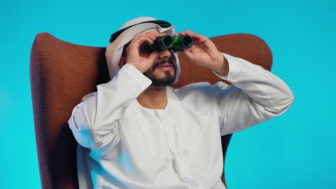 حب الاستكشاف والمغامرة،  استعمال المنظار للكشف عن الاشياء البعيدة وتقريبها,  رجل عربي خليجي اماراتي يرتدي الكندورة والغتره يجلس على كرسي مريح يحمل بيده المنظار ثنائي العينين وينظر الى شيء ما، إيماءات الاستمتاع والسرور