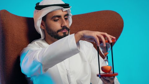 Realizing the value of precious moments in life, planning to achieve future goals, waiting for something eagerly, an Arab Gulf Emirati man wearing a kandura and ghutrah sitting on a chair holding an hourglass, observing the passage of time with gestures of happiness and joy.