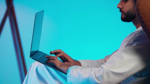 Making video calls with family and friends, remote work meetings, a side shot of an Emirati Gulf Arab man wearing a kandura and ghutrah with headphones, holding a laptop and looking at it with gestures of happiness, using modern technologies.