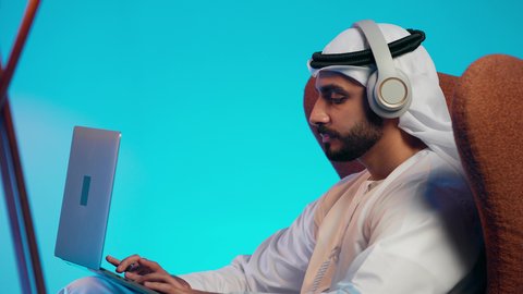 The use of modern technologies, making video calls with family and friends, remote work meetings, a side shot of an Arab Gulf Emirati man wearing a kandura and ghutrah, with headphones, holding a laptop and looking at it with gestures of happiness.