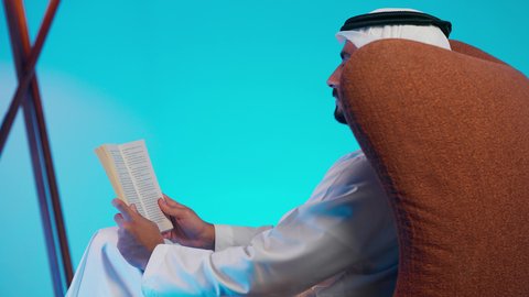 تمضية أوقات الفراغ بالتعلم والتثقف الذاتي, إيماءات الانتباه والتركيز ,  تصوير من الجانب لرجل عربي خليجي اماراتي يرتدي الكندورة والغترة يقوم بالقراءة بتمعن, مطالعة القصص التصويرية المسلية, تنمية مهارات القراءة , الاهتمام بتصفح الكتب المفيدة