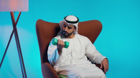 رجل عربي خليجي اماراتي يرتدي الكندورة والغتره  يقوم برفع الأوزان الحديدية الثقيلة، ممارسة التمارين الرياضية مع الدمبل،تمارين رفع الأثقال (دمبلز)، المحافظة على الصحة واللياقة الجسدية
