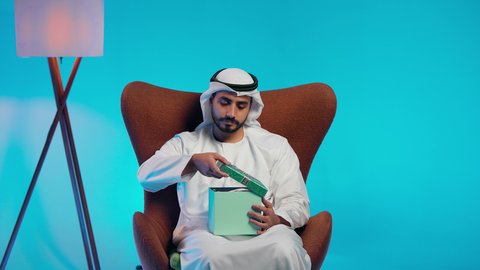تقديم الهدايا بالاعياد والمناسبات الخاصة، مفهوم الشكر والامتنان، التعبير عن الحب والتقدير والاحترام، رجل عربي خليجي اماراتي يرتدي الكندورة والغتره يجلس على كرسي مريح يحمل بيديه صندوق هدية, ايماءات السرور والسعادة