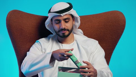 مفهوم الشكر والامتنان، تقديم الهدايا بالاعياد والمناسبات الخاصة، التعبير عن الحب والتقدير والاحترام، رجل عربي خليجي اماراتي يرتدي الكندورة والغتره يجلس على كرسي مريح يحمل بيديه صندوق هدية, ايماءات السرور والسعادة