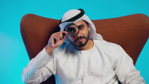 الفحص والاستكشاف والرؤية عن قرب، مفهوم توضيح الصورة، ايماءات السعادة والسرور،رجل عربي خليجي اماراتي يرتدي الكندورة والغترة ينظر من خلال العدسة المكبرة، التدقيق والتركيز باستخدام الأدوات البصرية