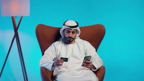 التسويق اونلاين عبر الانترنت ، الدفع عبر بطاقة الائتمان ، رجل عربي خليجي اماراتي يرتدي الكندورة والغتره يمسك الجوال والبطاقة بيده ،دمج التقنيات الحديثة فى الحياة اليومية