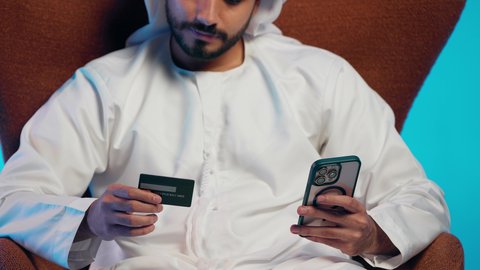 الدفع عبر بطاقة الائتمان ، التسويق اونلاين عبر الانترنت ، رجل عربي خليجي اماراتي يرتدي الكندورة والغتره يمسك الجوال والبطاقة بيده ،دمج التقنيات الحديثة فى الحياة اليومية