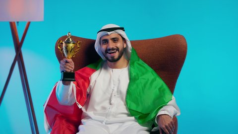 مشاهدة ومتابعة مباراة كرة القدم،  رجل عربي خليجي اماراتي يرتدي الكندورة والغتره والعلم الاماراتي يجلس على كرسي مريح يحمل كأس المنتخب، الحماس والتفاعل مع المنتخب، إيماءات الشغف والتشجيع للفريق، أنديه رياضية مختلفة