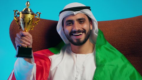 إيماءات الشغف والتشجيع للفريق، الحماس والتفاعل مع المنتخب،مشاهدة ومتابعة مباراة كرة القدم،  رجل عربي خليجي اماراتي يرتدي الكندورة والغتره والعلم الاماراتي يجلس على كرسي مريح يحمل كأس المنتخب، أنديه رياضية مختلفة