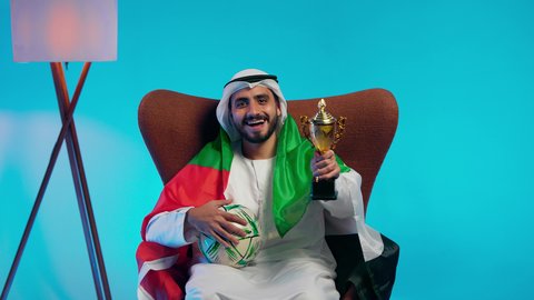 الحماس والتفاعل مع المنتخب، إيماءات الشغف والتشجيع للفريق، مشاهدة ومتابعة مباراة كرة القدم،  رجل عربي خليجي اماراتي يرتدي الكندورة والغتره والعلم الاماراتي يجلس على كرسي مريح يحمل كأس المنتخب، أنديه رياضية مختلفة