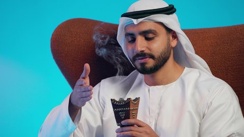 The use of oud wood during holidays and occasions, Emirati customs and traditions, an Arab Gulf Emirati man wearing a kandura and ghutrah sitting on a comfortable chair using a incense burner, attention to external appearance and elegance, perfuming and scenting with incense.