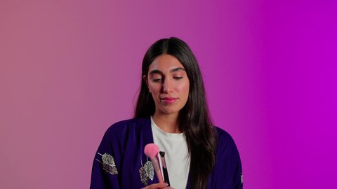 العناية بالبشرة  بواسطة المستحضرات, مفهوم التزين والتجمل, الاهتمام بالمظهر الخارجي، بورتريه مقرب لامرأة عربية خليجية  سعودية ترتدي الزي الكاجوال تستعمل فرشاة أحمر الخدود, خلفية ملونه