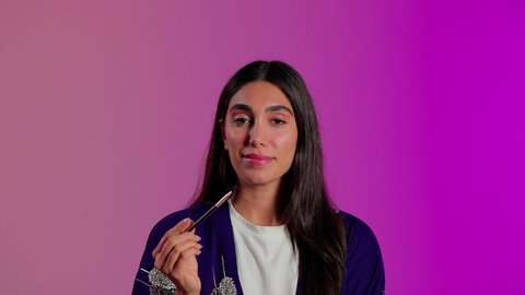 Promoting beauty products and cosmetics, the concept of adornment and beautification, the use of cosmetic products, caring for beauty and appearance, a close-up portrait of a Saudi Gulf Arab woman wearing casual attire using an eyeshadow brush, colorful background.