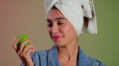 الحرص على تناول أطعمة مغذية ومتوازنة، أهمية وفائدة الفواكه الطازجة للبشرة، فطور وسناك لذيذ, بورتريه مقرب لامرأة عربية خليجية سعودية ترتدي الزي الكاجوال تضع منشفة بيضاء على رأسها تمسك تفاحة خضراء، خلفية ملونه