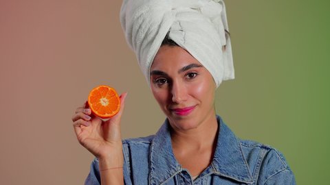 Delicious breakfast and snack, meeting the body's nutritional needs, a close-up portrait of a Saudi Gulf Arab woman wearing casual attire with a white towel on her head, holding slices of orange in her hands and placing them in front of her eyes, following a healthy diet, the importance of fresh fruits for the skin, colorful background.