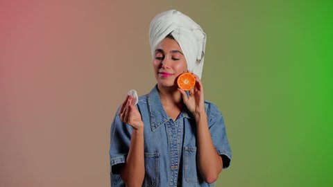 علاج تجميلي طبيعي للحفاظ على النضارة، بورتريه لامرأة عربية خليجية سعودية ترتدي الزي الكاجوال تضع منشفة بيضاء على رأسها تمسك بيديها شريحه برتقال قطنه تقوم بمسح وجهها، إزالة المكياج باستخدام ضمادات قطنية، الاهتمام بالمظهر الخارجي، خلفية ملونه