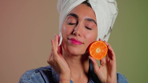 العناية بالبشرة بمكونات طبيعية آمنة، بورتريه مقرب لامرأة عربية خليجية سعودية  تضع منشفة بيضاء على رأسها تمسك بيديها شريحه برتقال قطنه تقوم بمسح وجهها، إزالة المكياج باستخدام ضمادات قطنية، الاهتمام بالمظهر الخارجي، خلفية ملونه
