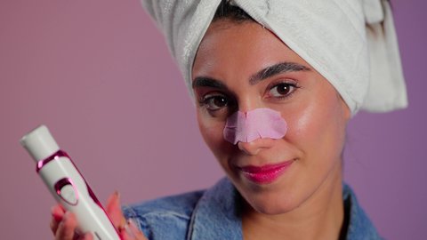 إستخدام أداة شفط لحب الشباب،  روتين العناية بالبشرة، ماسك تنظيف المسام، بورتريه مقرب لمرأة عربية خليجية سعودية ترتدي منشفة الرأِس تضع لصقات الأنف للتخلص من الرؤوس السوداء والجلد الميت، مفهوم العناية والإهتمام بنضارة وتحسين البشرة ، خلفية ملونه