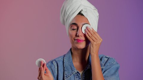 إزالة المكياج باستخدام ضمادات قطنية، الاهتمام بالمظهر الخارجي، مفهوم العناية بنضارة ونظافة البشرة، بورتريه مقرب لامرأة عربية خليجية سعودية تضع منشفة بيضاء على رأسها تمسك بيديها قطنتين تقوم بمسح وجهها، خلفية ملونة