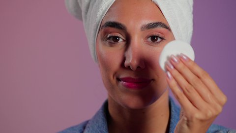 الاهتمام بالمظهر الخارجي، مفهوم العناية بنضارة ونظافة البشرة، بورتريه مقرب لامرأة عربية خليجية سعودية تضع منشفة بيضاء على رأسها تمسك بيديها قطنتين تقوم بمسح وجهها، إزالة المكياج باستخدام ضمادات قطنية، خلفية ملونة