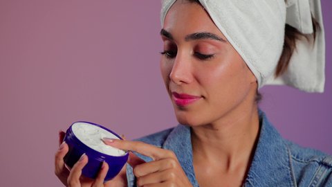 استخدام كريمات و مكونات تجميلية، مفهوم العناية بنضارة وتحسين البشرة، تدليك و ترطيب الوجه، الاهتمام بالمظهر الخارجي، بورتريه مقرب لامرأة عربية خليجية سعودية ترتدي  منشفة بيضاء تضع كريم مرطب، خلفية ملونه