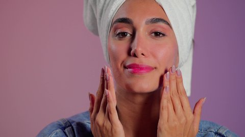 تدليك و ترطيب الوجه، الاهتمام بالمظهر الخارجي، مفهوم العناية بنضارة وتحسين البشرة، بورتريه مقرب لامرأة عربية خليجية سعودية ترتدي  منشفة بيضاء تضع كريم مرطب، استخدام كريمات و مكونات تجميلية، خلفية ملونه