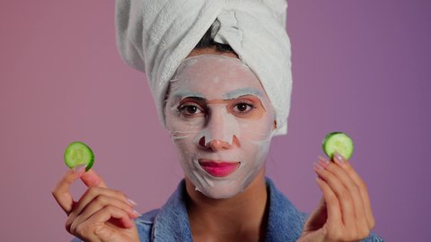 Skincare routine to maintain its freshness, a close-up portrait of a Saudi Gulf Arab woman wearing a head towel applying a facial mask with a cucumber mask for the eyes, using cosmetic ingredients, colorful background.