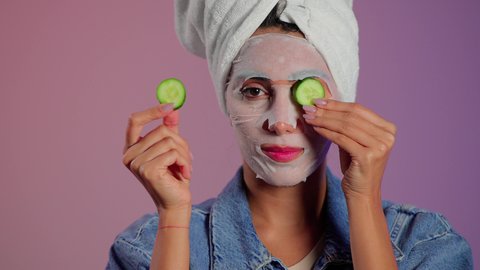 الاهتمام بالبشرة والحفاظ على نضارتها، بورتريه مقرب لمرأة عربية خليجية سعودية ترتدي منشفة الرأس تضع القناع التجميلي على الوجه مع قناع الخيار للعيون، مفهوم العناية والإهتمام بنضارة وتحسين البشرة، إستخدام مكونات تجميلية، خلفية ملونه