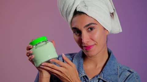 تنظيف البشرة وترطيبها لتحسين مظهرها، الاهتمام بالأناقة والمظهر الخارجي، بورتريه مقرب لامرأة عربية خليجية سعودية ترتدي منشفة بيضاء على رأسها تضع كريم الترطيب على وجهها، استخدام مستحضر ومكون تجميلي، خلفية ملونه