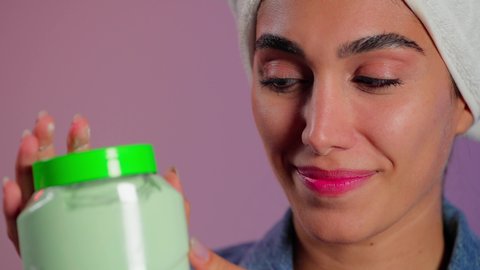 الاهتمام بالأناقة والمظهر الخارجي، مفهوم العناية بنضارة وتحسين البشرة، بورتريه مقرب لامرأة عربية خليجية سعودية ترتدي منشفة بيضاء على رأسها تضع كريم الترطيب على وجهها، استخدام مستحضر ومكون تجميلي، خلفية ملونه