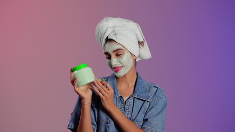 Using cosmetic ingredients and products, the concept of care and attention to women's appearance, steps for cleansing and exfoliating the skin, a close-up portrait of a Saudi Gulf Arab woman wearing a white towel holding cream in her hands and placing slices of cucumber on her face, colorful background.