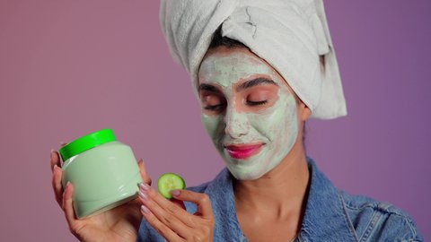 مفهوم العناية والاهتمام بالمظهر الخارجي عند النساء، خطوات تنظيف وتقشير البشرة، استخدام مكونات و مستحضرات تجميلية، بورتريه مقرب لامرأة عربية خليجية سعودية ترتدي منشفة بيضاء  تمسك بيديها الكريم وتقوم بوضع قطع من الخيار على وجهها، خلفية ملونه