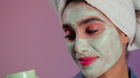 رعاية البشرة للحفاظ على حيويتها ونضارتها، بورترية مقرب لامراة عربيه خليجيه سعودية ترتدي منشفة الرأس تمسك علبه كريم تقوم بوضعه على وجهها ، إستخدام مكونات تجميلية، خلفية ملونه