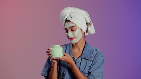 روتين العناية بالبشرة، بورترية لامراة عربيه خليجيه سعودية ترتدي الزي الكاجوال و منشفة الرأس تمسك علبه كريم تقوم بوضعه على وجهها، مفهوم العناية والإهتمام بنضارة وتحسين الجلد ، إستخدام مكونات تجميلية، خلفية ملونه