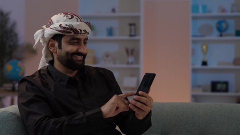 A Yemeni Arab man wearing a shirt and a sarong with a turban sits and holds a mobile phone, showing gestures of happiness while communicating with friends and relatives, exchanging conversations and good news, representing authentic Arab masculinity, maintaining traditional attire, using a modern and advanced technological device, embodying the concept of dignity and elegance.
