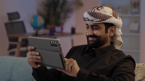 رجل عربي يمني يرتدي القميص والازار مع العمامة يمسك بيده جهاز تابلت بإيماءات الجدية والتركيز، استعمال التكنولوجيا الحديثة، تصفح تطبيقات التواصل الاجتماعي، استخدام الجهاز اللوحي في إنجاز مهام العمل، الدردشة والتحدث مع الاصدقاء عبر الانترنت