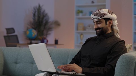 رجل عربي يمني يرتدي القميص والازار مع العمامة  يجلس على اريكة مريحة يقوم بالعمل على الحاسب المحمول ،تعابير السرور والسعادة، إدارة شؤون الأعمال وتنظيمها، كتابة وإدخال المعلومات عبر الكيبورد، استخدام التقنيات الحديثة المتطورة، عقد إجتماع عمل عبر الإنترنت