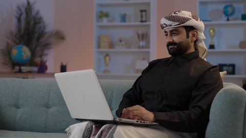 A Yemeni Arab man wearing a shirt and a sarong with a turban is sitting on a comfortable sofa working on a laptop, utilizing modern devices in work, managing and organizing business affairs, typing and entering information via the keyboard, using advanced modern technologies, and holding a business meeting online.