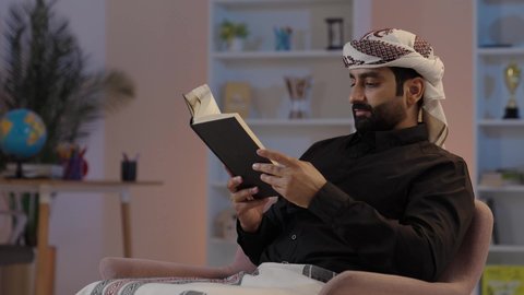 A Yemeni Arab man wearing a shirt and a sarong with a turban sits holding a book, preserving traditional attire, reading and being interested in old books, the concept of dignity and elegance, focusing on browsing useful books, engaging oneself in reading and education, the concept of contemplation and thinking, representing authentic Arab masculinity.