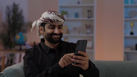 A Yemeni Arab man wearing a shirt and a sarong with a turban sits holding a mobile phone, engaging in online shopping and browsing programs, exchanging conversations and good news, showing gestures of happiness while connecting with friends and relatives, representing authentic Arab masculinity, maintaining traditional attire, and embodying the concept of dignity and elegance.