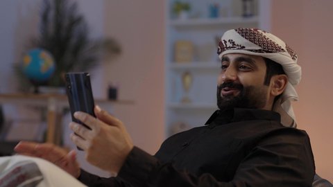 رجل عربي يمني يرتدي القميص والازار مع العمامة يجلس ويحمل الهاتف المحمول، السلام والتحية خلال مكالمة فيديو، تبادل الأحاديث و الأخبار الجيدة، إيماءات السعادة بالتواصل مع الأصدقاء والأقارب، تمثيل الرجولة العربية الأصيلة، المحافظة على اللباس التقليدي