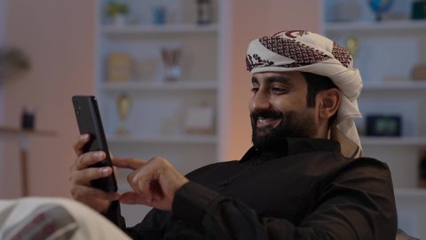 A Yemeni Arab man wearing a shirt and a sarong with a turban sits holding a mobile phone, sharing news and good conversations, exchanging good talks and news, showing gestures of happiness in connecting with friends and relatives, representing authentic Arab masculinity, and preserving traditional attire.