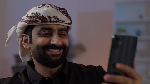 رجل عربي يمني يرتدي القميص والازار  مع العمامة يجلس ويحمل الهاتف المحمول، تصفح الإنترنت ، التواصل مع الأصدقاء، تبادل الأحاديث و الأخبار الجيدة، إيماءات السعادة بالتواصل مع الأصدقاء والأقارب، تمثيل الرجولة العربية الأصيلة، المحافظة على اللباس التقليدي