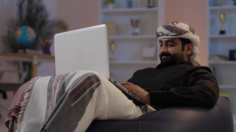 رجل عربي يمني يرتدي القميص والازار مع العمامة يجلس على كرسي مريح يقوم بالعمل على الحاسب المحمولإدارة شؤون الأعمال وتنظيمها، كتابة وإدخال المعلومات عبر الكيبورد، دمج التكنولوجيا في الحياة العملية، استخدام التقنيات الحديثة المتطورة، عقد إجتماع عمل عبر الإنترنت،