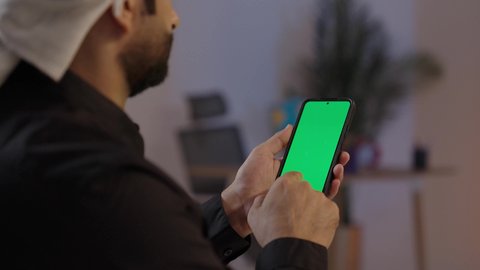 Close-up shot of a Yemeni Arab man wearing a shirt and a sarong with a turban and a jambiya, standing and holding a phone, with a green phone screen background, the concept of body language and facial expressions, representing authentic Arab masculinity, modern technological devices in daily life, the concept of dignity and elegance, beige background.