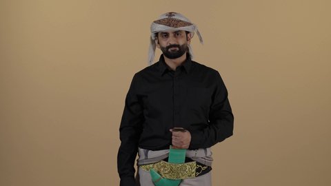 رجل  عربي يمني مبتسم يرتدي القميص الازار مع العمامة والجنبية يقوم بوضع يده على الجنبية و يحريك يديه، ايماءات الحدة والجدية، اشارة توقف لاتكمل، مفهوم العلاقات الاجتماعية، المحافظة على اللباس التقليدي، مفهوم الهيبة والرجولة، خلفية بيج