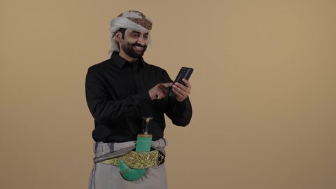 بورتريه رجل عربي يمني يرتدي القميص والازار مع العمامة  والجنبية يقف ويحمل هاتف، الضحك خلال التواصل الاجتماعي والتصفح، مفهوم لغة الجسد وتعابيرالوجه، تمثيل الرجولة العربية الأصيلة، الاجهزة التقنية الحديثة في الحياة اليومية،  مفهوم الهيبة والاناقة، خلفية بيج